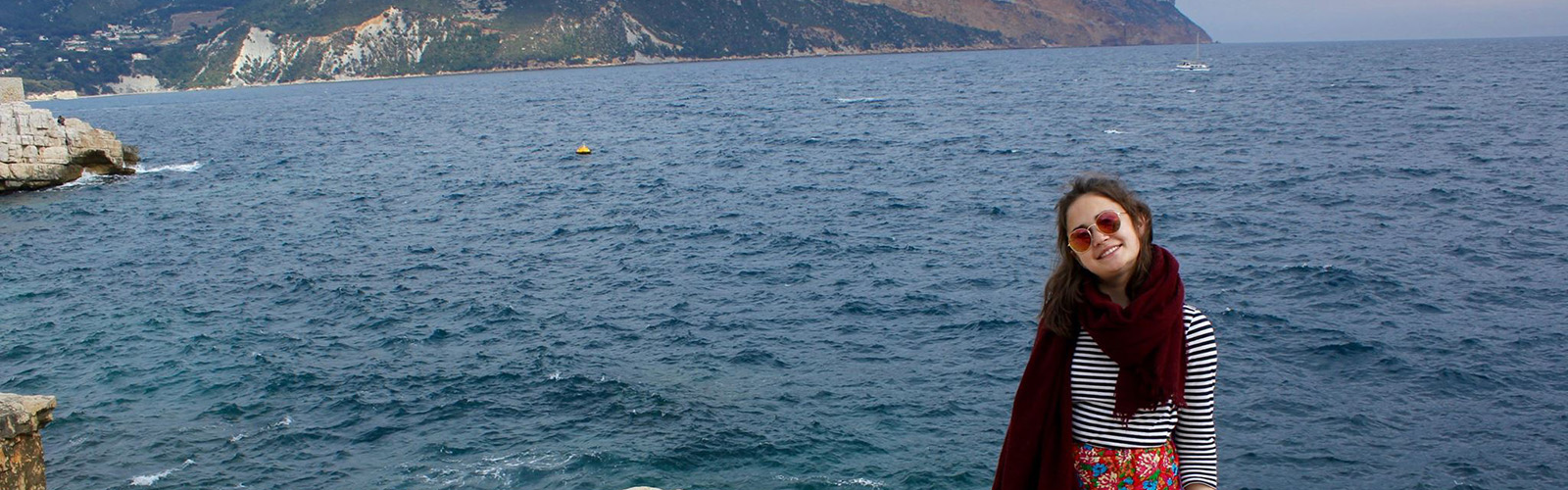 A student is posed in front of the ocean. Waves are crashing onto the rocky cliffs around them. They are smiling at the camera.